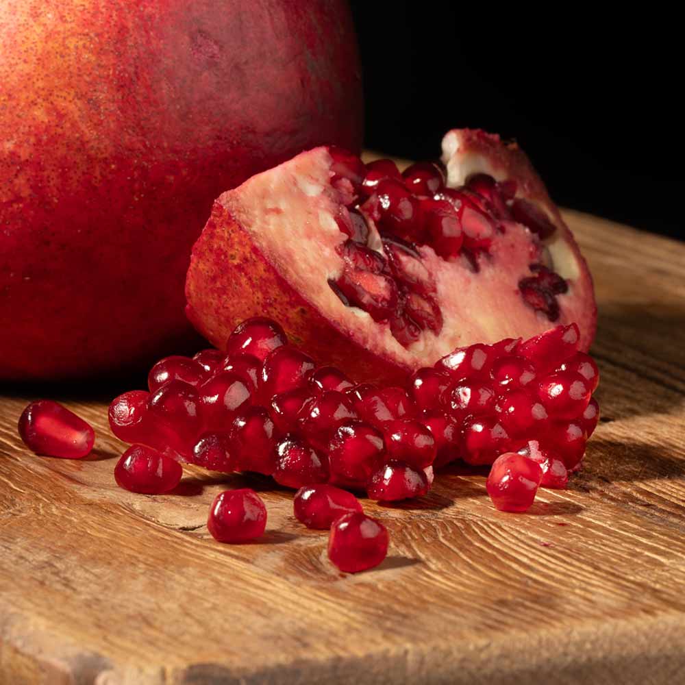 A close-up of a whole and halved pomegranate with bright red arils scattered on a wooden surface against a dark background.