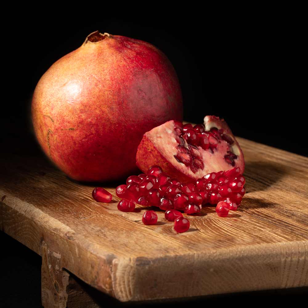 A whole pomegranate and a cut half showing red arils, with scattered pomegranate seeds on a rustic wooden board against a black background.