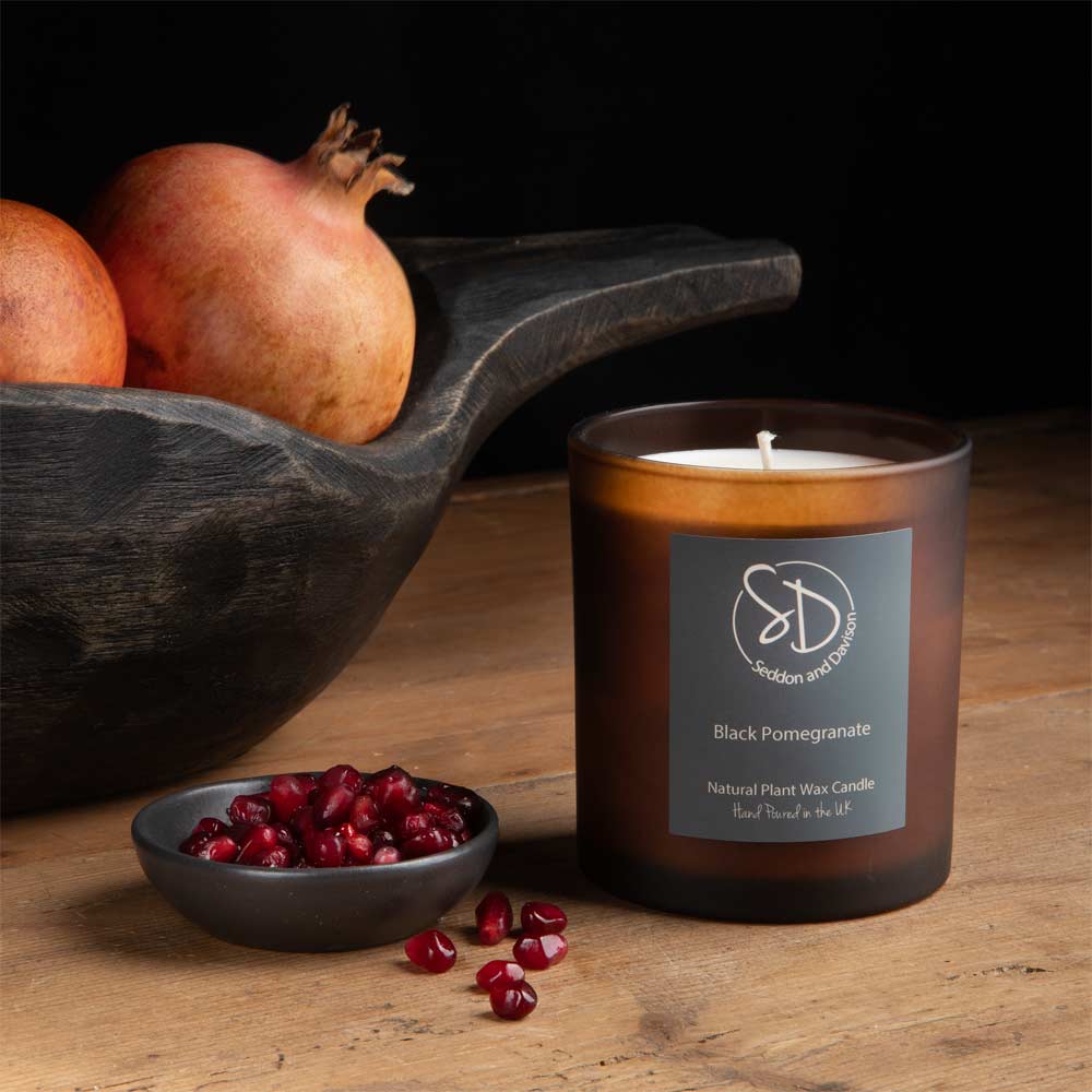 A brown amber glass jar candle labeled 'Black Pomegranate' by Seddon and Davison, styled on a wooden table with pomegranate seeds and whole pomegranates in a dark wooden bowl.