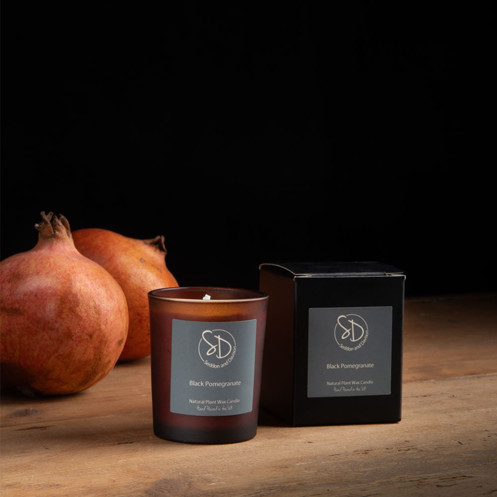 An amber glass votive candle  labelled 'Black Pomegranate' stands next to its black box on a wooden surface, with two whole pomegranates behind it against a black background.