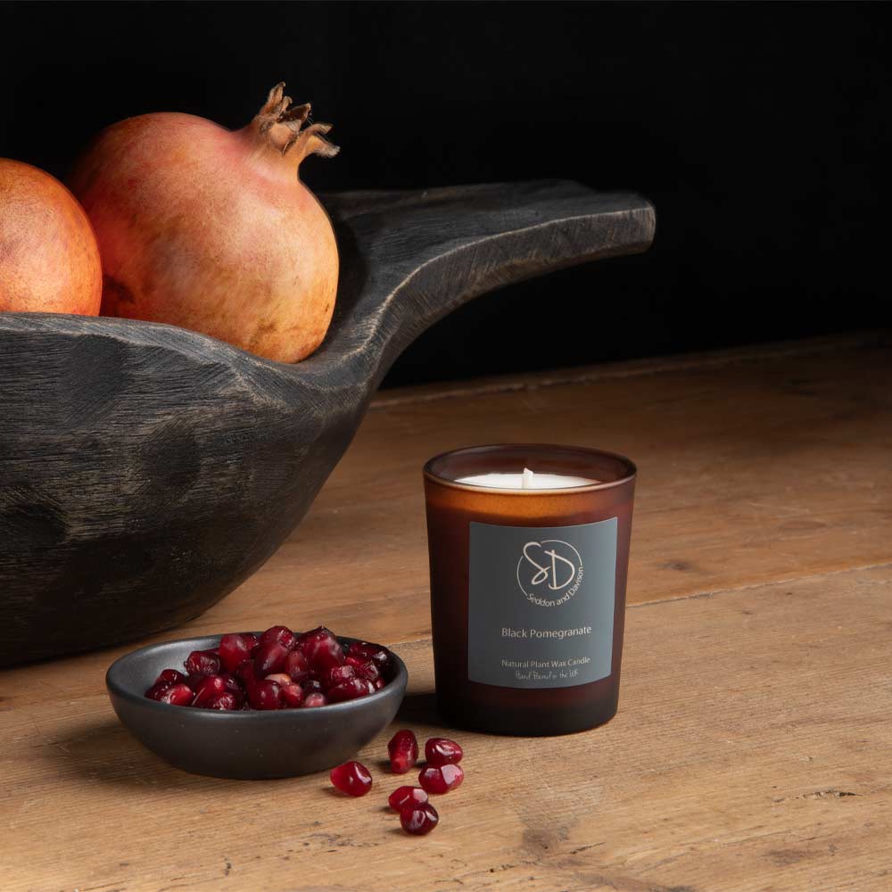 A dark amber glass Votive candle labeled 'Black Pomegranate' by Seddon and Davison, styled on a wooden table with a bowl of pomegranate seeds and whole pomegranates in a dark wooden bowl.