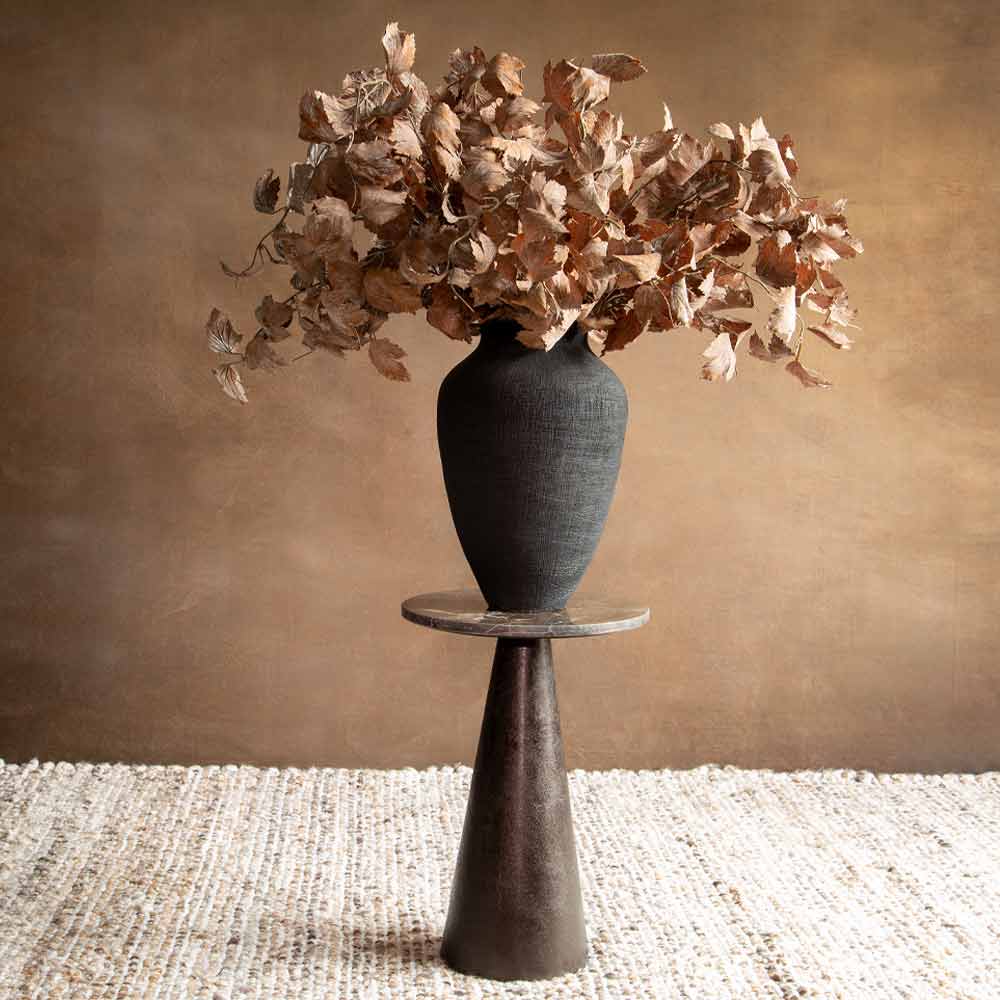 A Faux leaf arrangement in a black textured ceramic vase, displayed on a round marble topped pedestal against a warm brown wall.