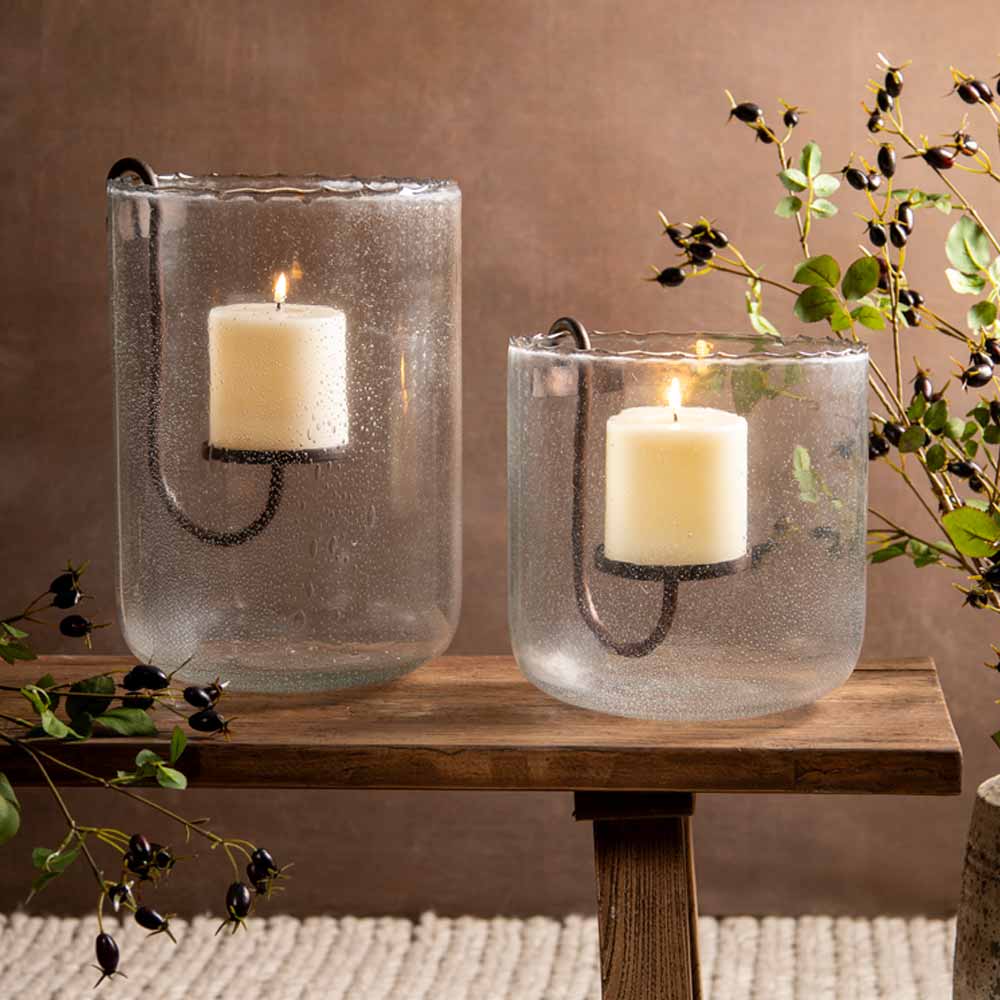 Two clear glass candle holders with a textured, bubbly finish, each containing a lit cream pillar candle on a  metal U-shaped bracket. The holders are displayed on a rustic wooden bench against a warm brown background, decorated with green and black rose hip branches.