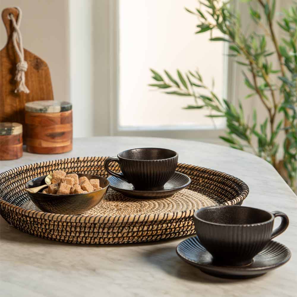 Black ribbed ceramic cups and saucers displayed on a woven tray on  a marble counter top, with a bowl made of horn holding sugar cubes. The scene is styled with wooden containers, a cutting board, and a potted olive tree in a bright, natural setting.