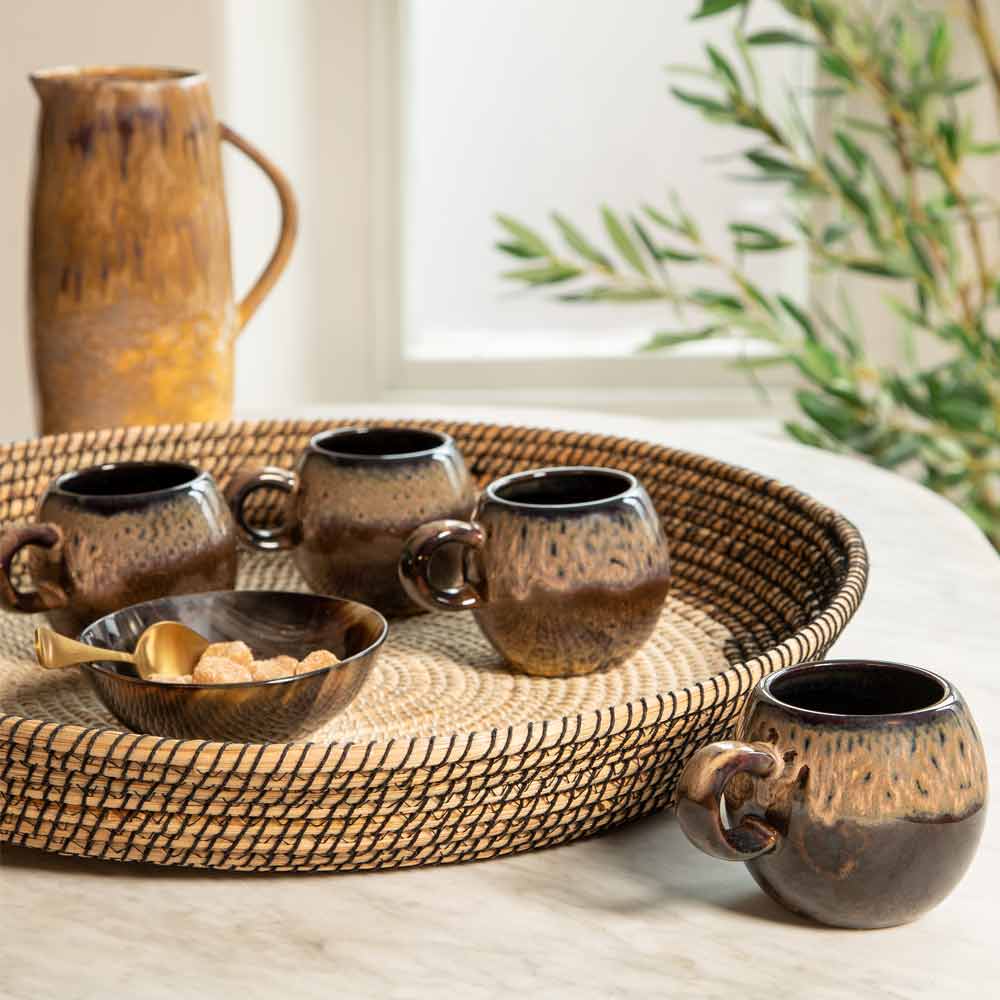 warm chestnut and brown stotoneware cups displayed on a woven tray on a marble counter top, with a bowl made of horn holding the sugar cubes. The scene is styled with a Jug and a potted olive tree in a bright, natural setting.