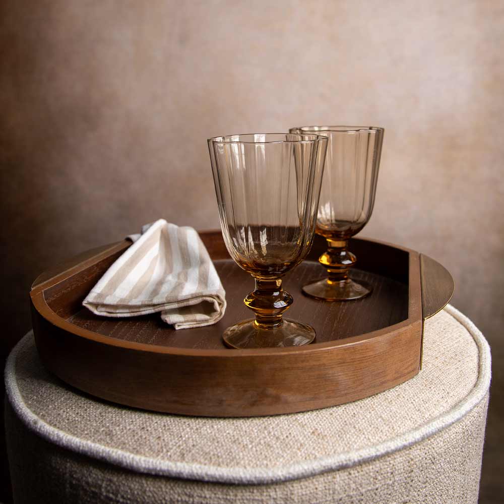 A round dark wood serving tray holding two amber-tinted stemware glasses and a folded striped cloth napkin, set on a textured beige upholstered stool against a warm neutral wall.