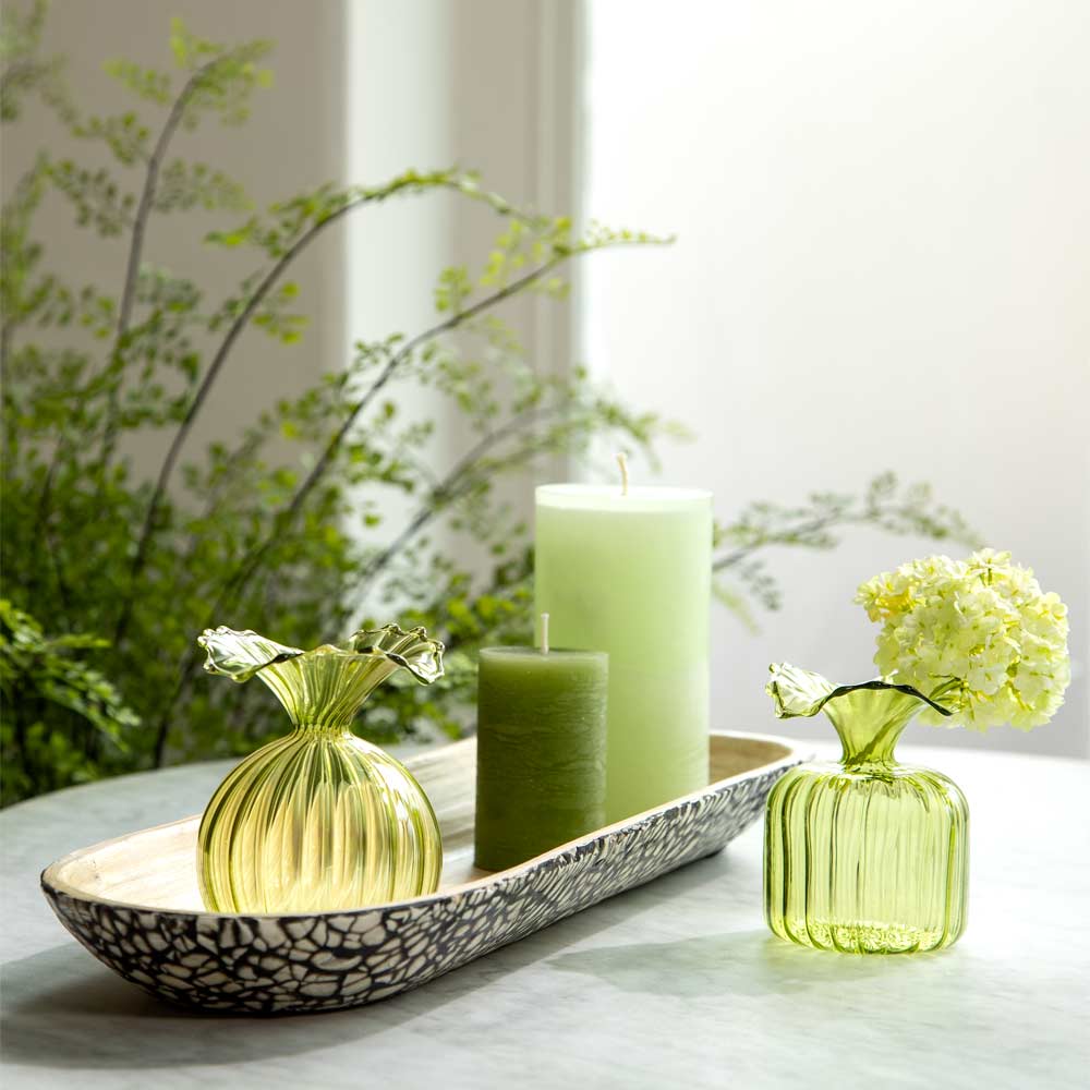 Decorative green glass vase and two green pillar candles arranged on a patterned tray and beside a small vase with pale yellow flower on a light marble tabletop, with soft-focus greenery in the background.