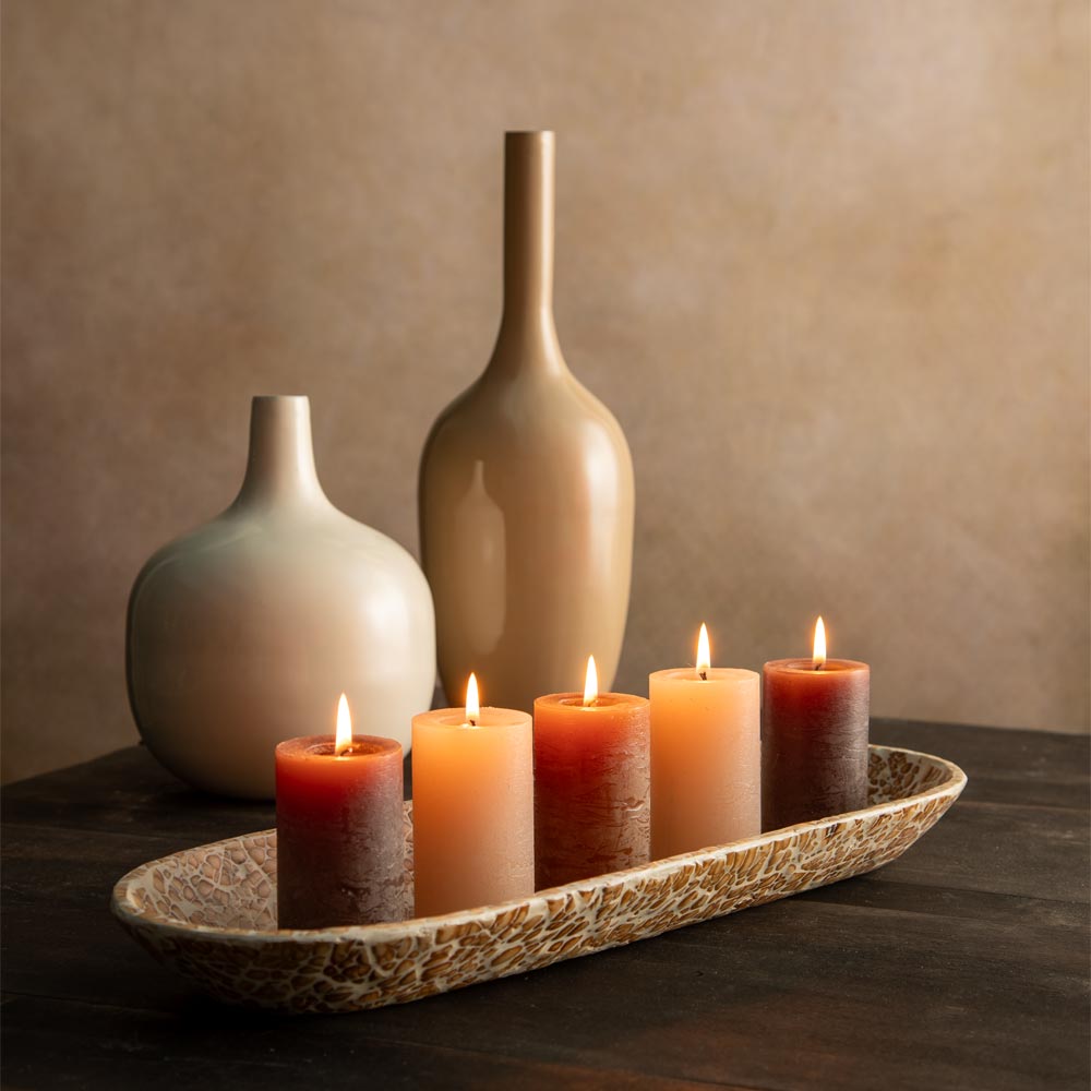 Five lit pillar candles in warm gradient shades from coffee, walnut  to nude-cream, arranged in a textured oval bamboo tray on a dark wooden table, with two matte powder coated vases in more neutral earthy shades in the background against a warm beige wall.