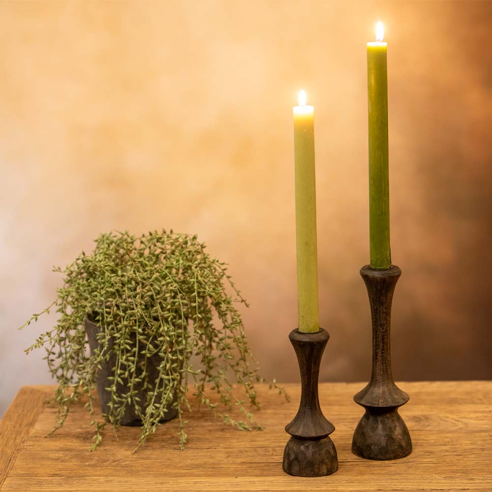 Two green candles in decorative holders on a wooden surface with a plant in the background.