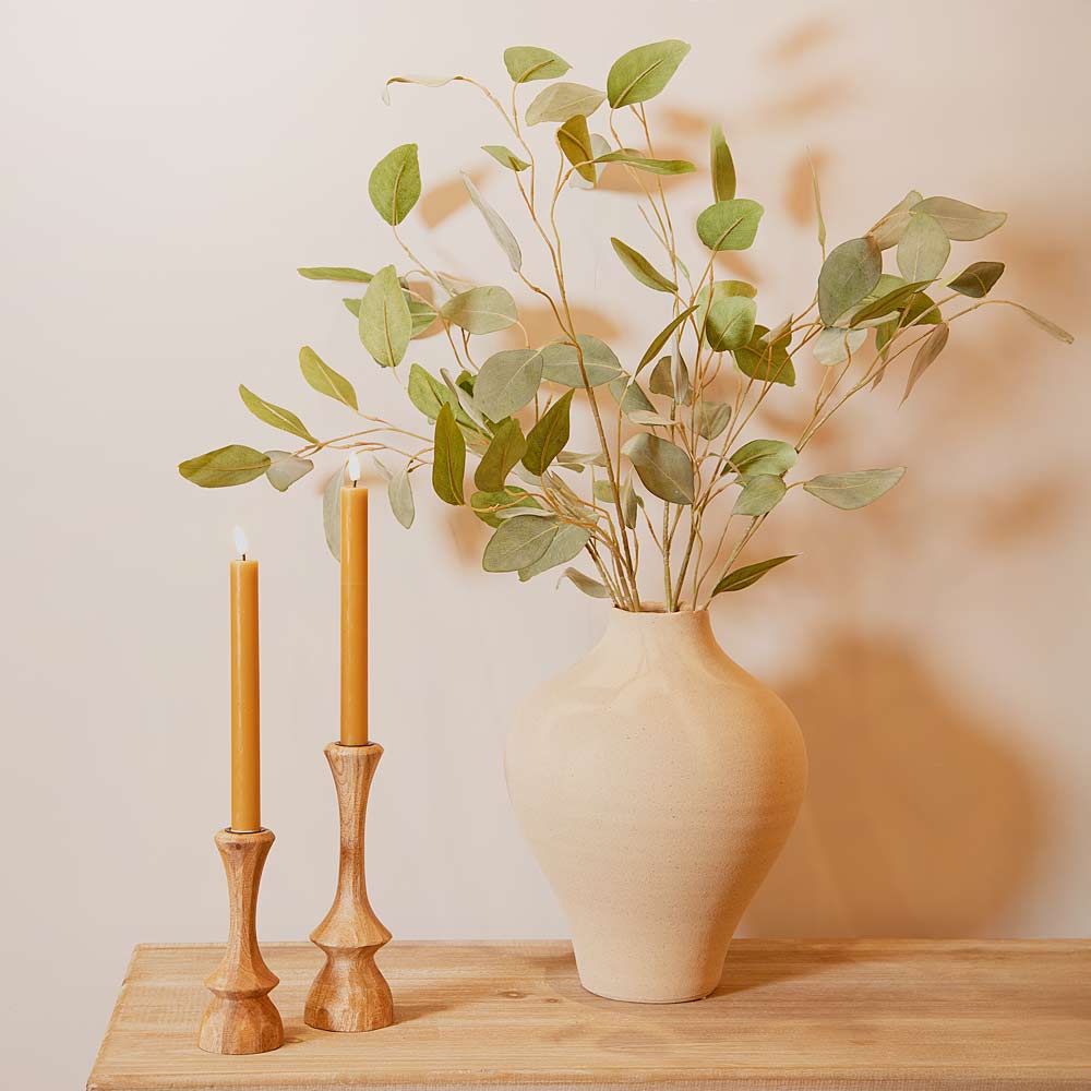 Vase with green leaves and two lit candles in the Cirilo Natural Wood Candlesticks on a wooden surface against a light background