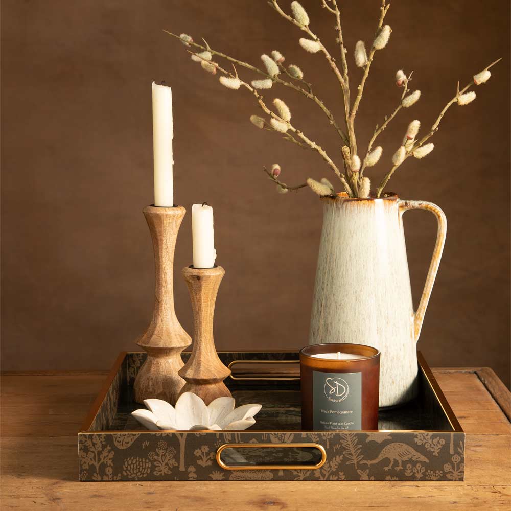 A styled tabletop arrangement featuring a decorative square tray with animal motifs and gold-toned handles, holding two wooden candlesticks with white taper candles, a ceramic pitcher with Faux Pussy Willow branches, and a amber glass   jar candle with a grey label.