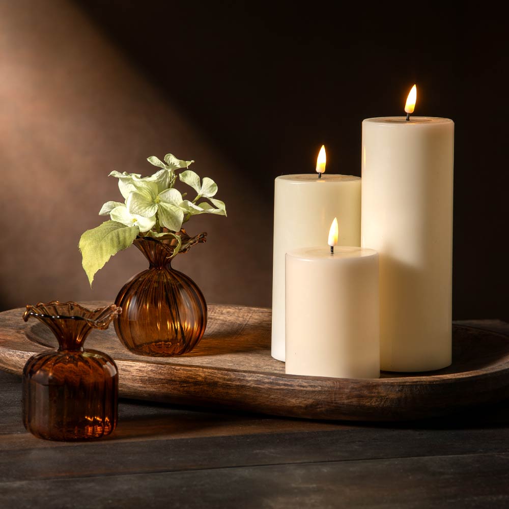Three cream pillar candles of different heights lit on a rustic wooden tray, with two small amber glass vases and pale green flowers and leaves beside them against a dark brown background.