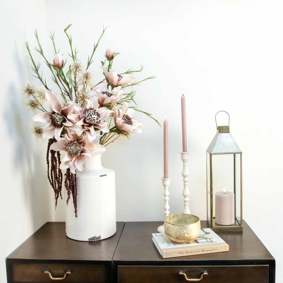 Ivory distressed bottle vase with faux flowers on a dark wood console, styled with pink candles and a lantern.