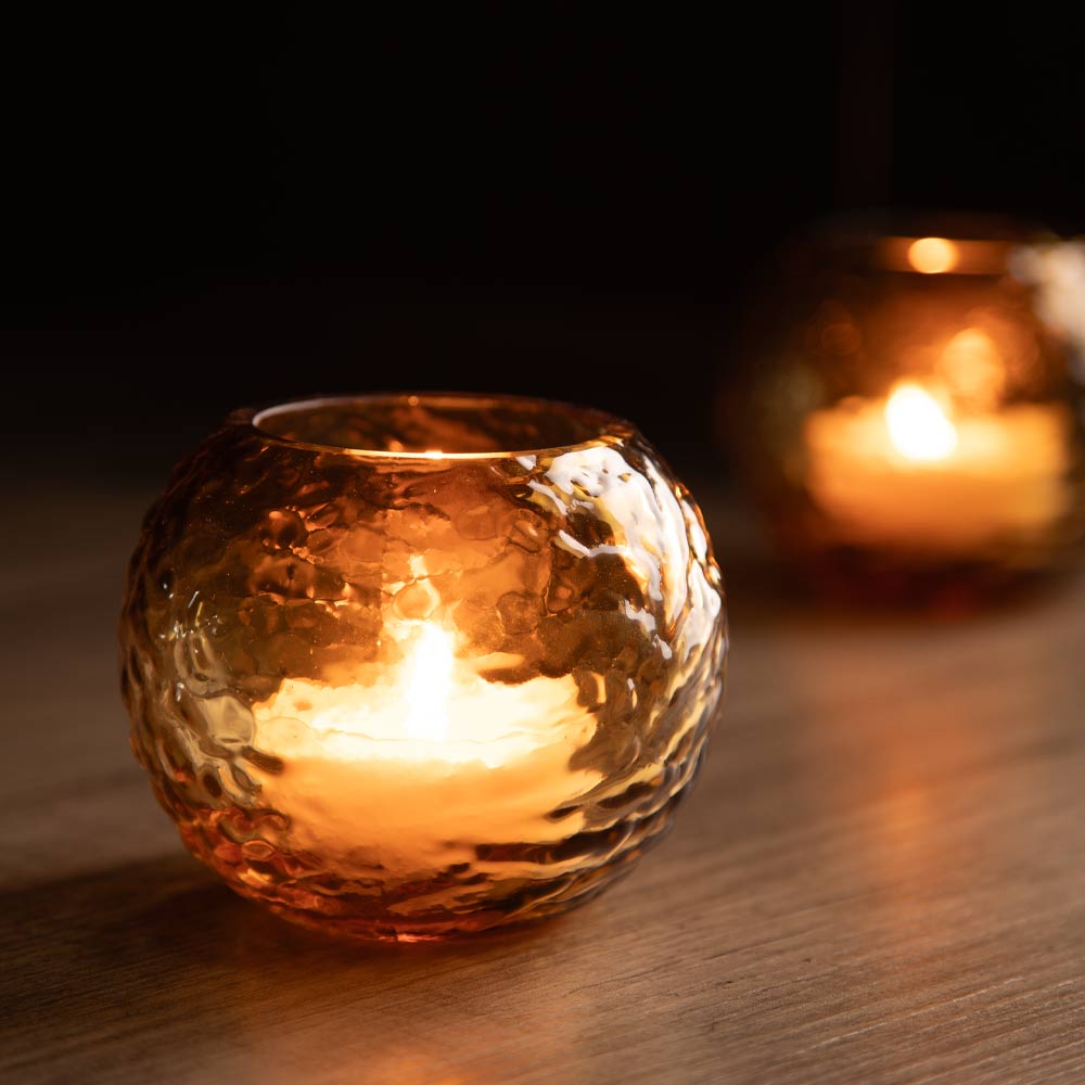 A small round amber glass candle holder with a hammered texture sits on a wooden surface with a lit candle, a second matching holder blurred in the background against a dark backdrop.
