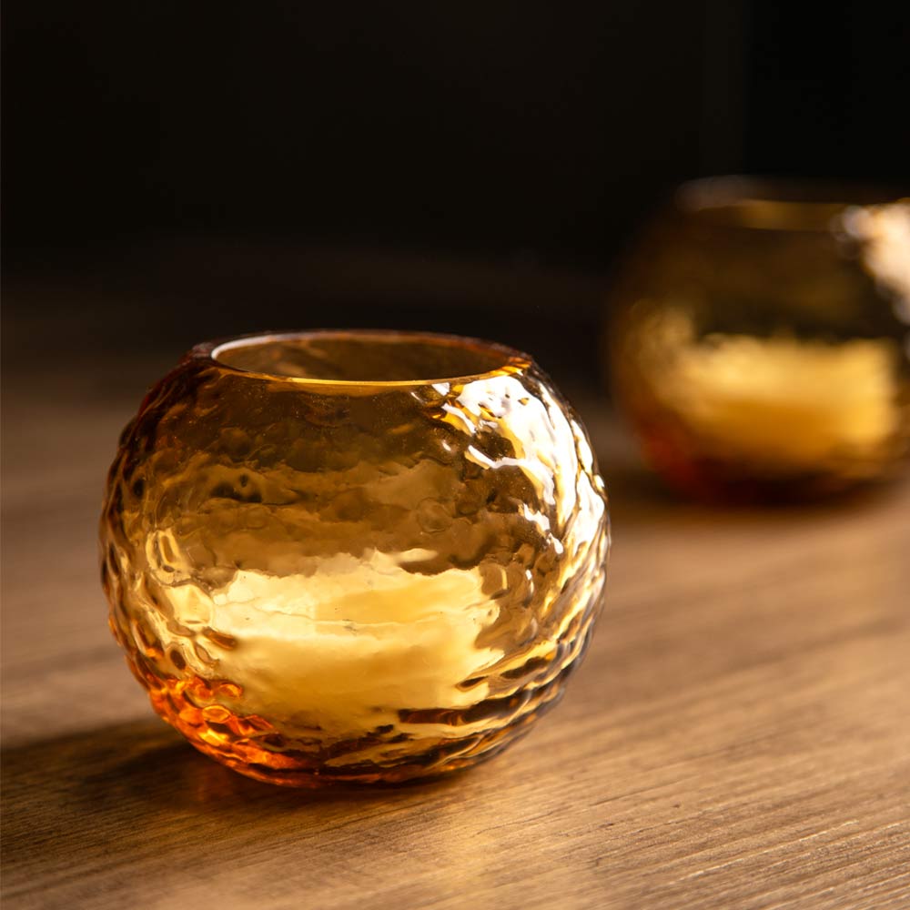A small round amber glass candle holder with a hammered texture sits on a wooden surface, with a second matching holder blurred in the background against a dark backdrop.