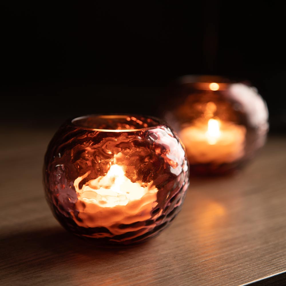 A close-up of a round plum glass candle holder with a hammered texture, containing a lit tea light candle that glows warmly against a dark background. A second matching holder is softly blurred in the background on a wooden surface.