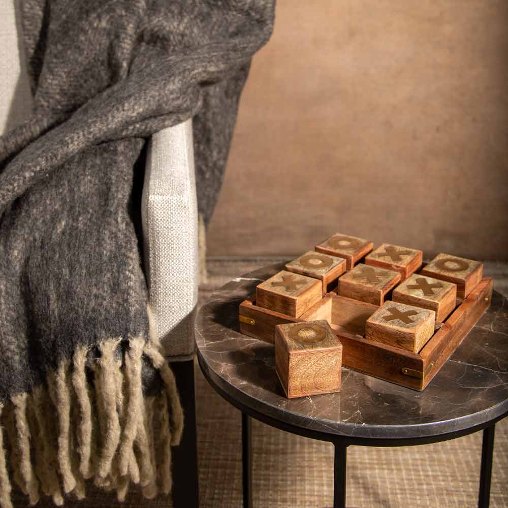 A set of wooden tic-tac-toe game blocks with brass inlay letters, displayed on a small round marble side table next to a cozy gray knit throw blanket draped over a chair.
