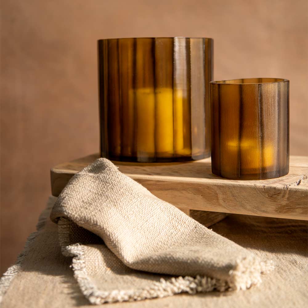Two amber glass candle holders, one tall and one short, displayed on a wooden tray with a beige linen cloth in the foreground against a warm brown background.
