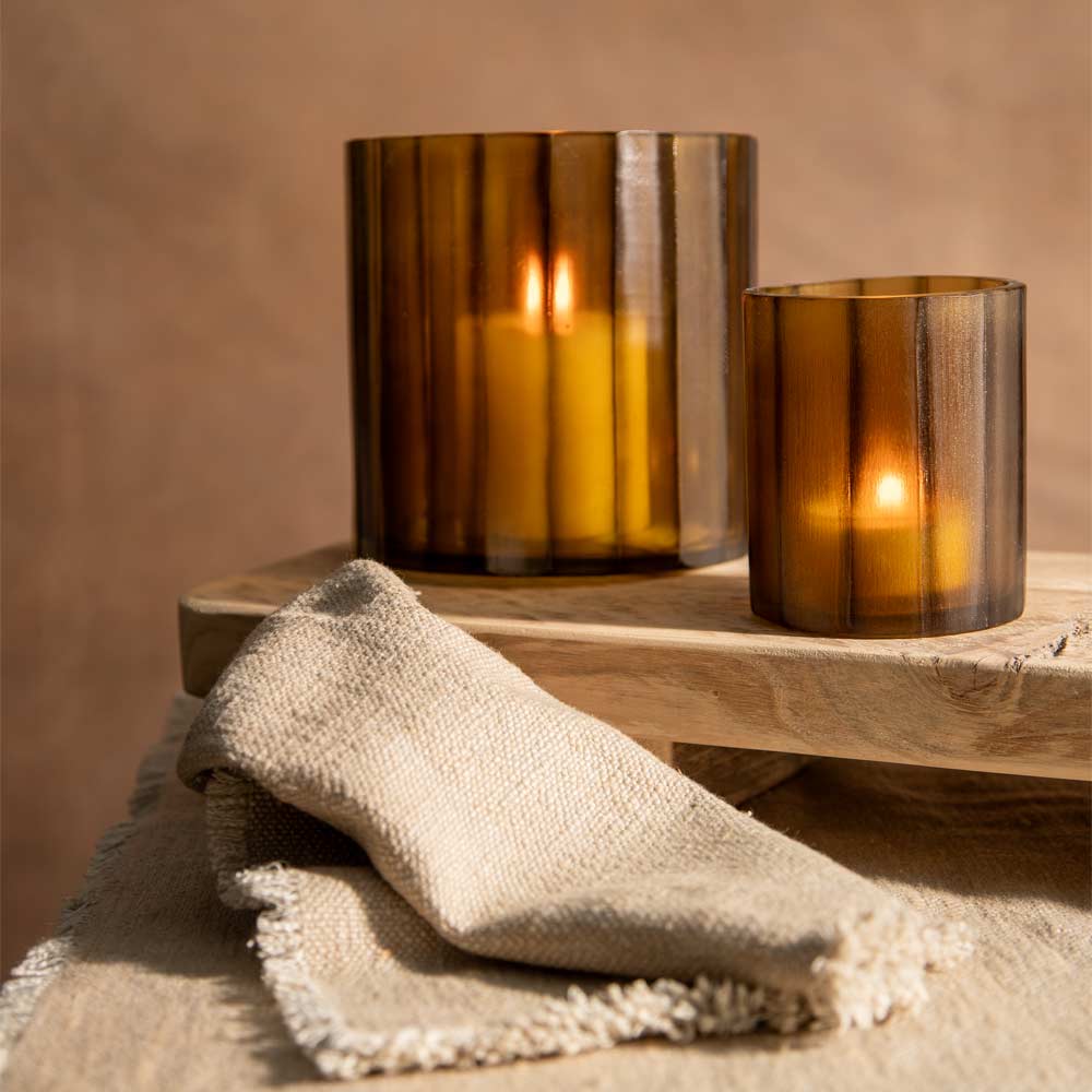 Two amber glass candle holders, one tall and one short, each containing a lit candle, styled on a wooden block with a beige linen cloth in the foreground against a warm beige background.