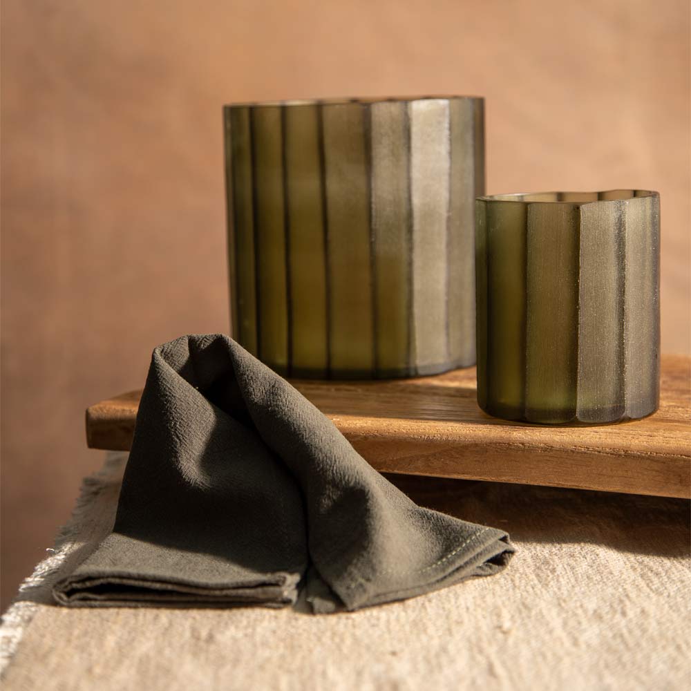 Two ribbed glass candle holders in a muted olive green and grey colour, styled on a wooden tray next to a dark cloth napkin against a warm beige background.