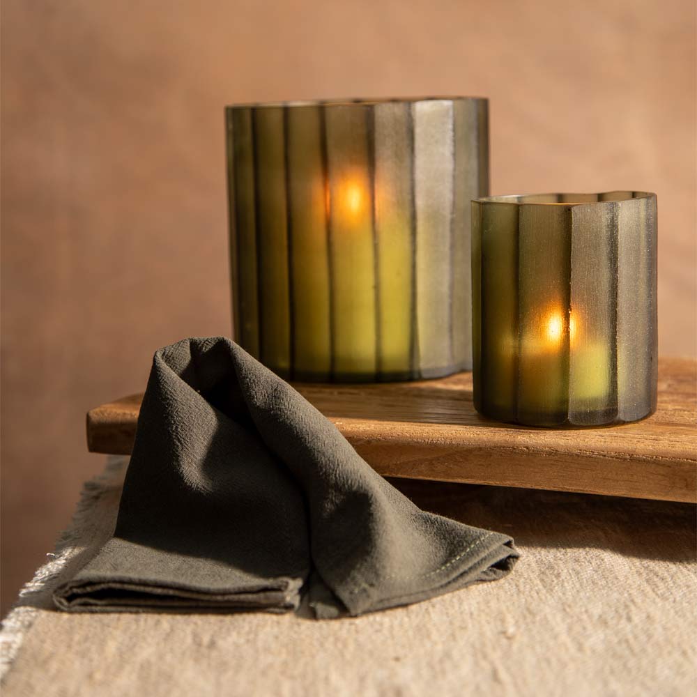 Two ribbed glass candle holders with a smoky olive-green finish, each containing a glowing lit candle, displayed on a wooden tray with a folded dark  cloth napkin in the foreground.