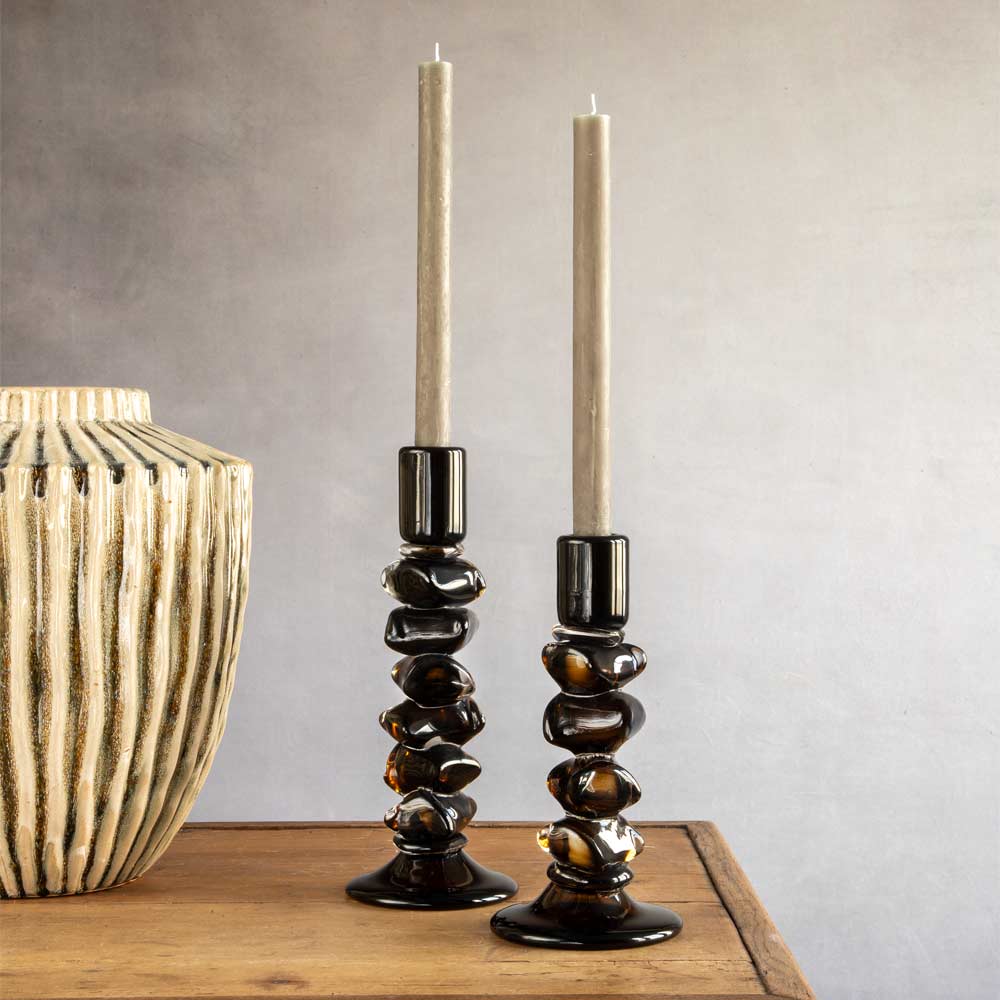 Two dark olive and black glass candlestick holders with twisted, organic shapes, holding Linen Dinner candles, displayed on a wooden table next to a ribbed ceramic vase against a neutral gray wall.