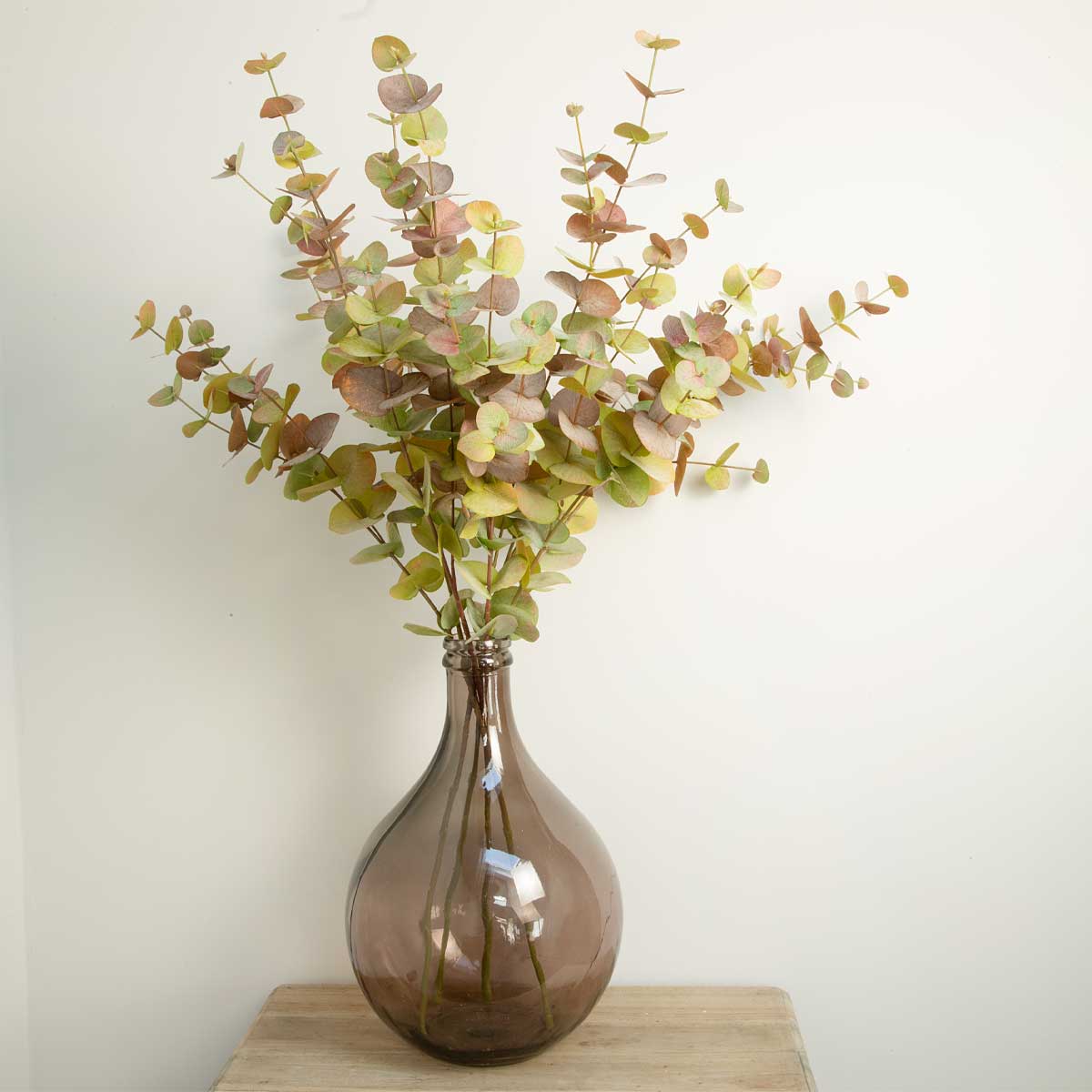 Artificial Tall Eucalyptus Autumn Spray in a tall, brown, glass vase, placed on a wooden surface against a white background.
