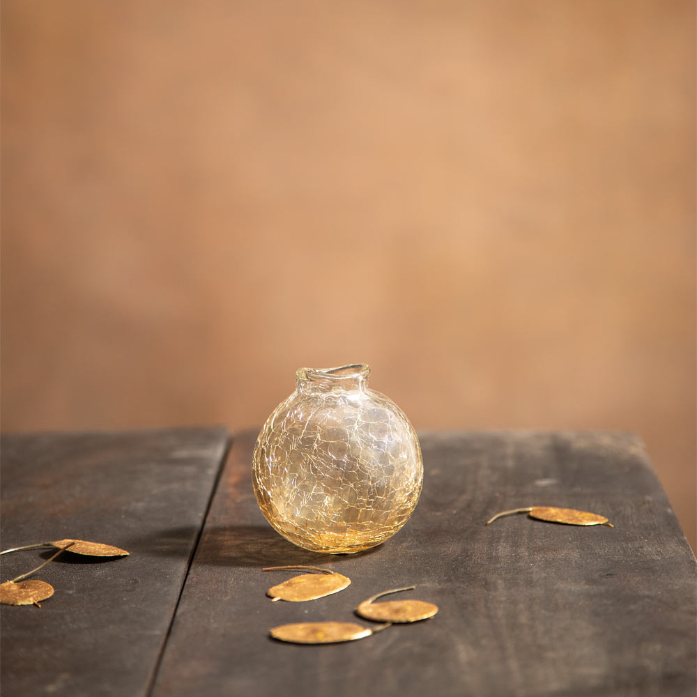 A small clear glass vase with a textured, crackled surface and a short neck, sitting on a dark wooden table with scattered dried leaves against a warm brown background.