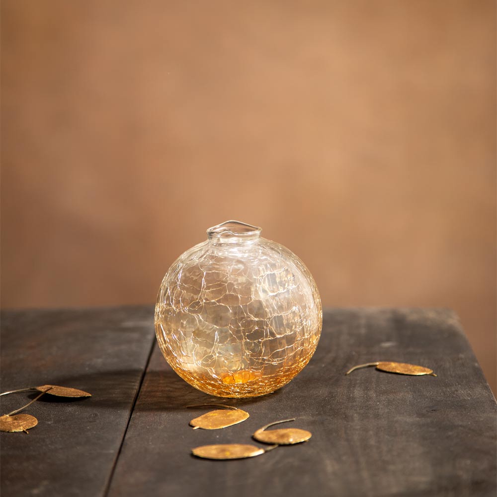 A small round clear glass vase with a textured crackle pattern and a narrow neck, sitting on a dark wooden surface with scattered gold-toned leaves against a warm brown background.