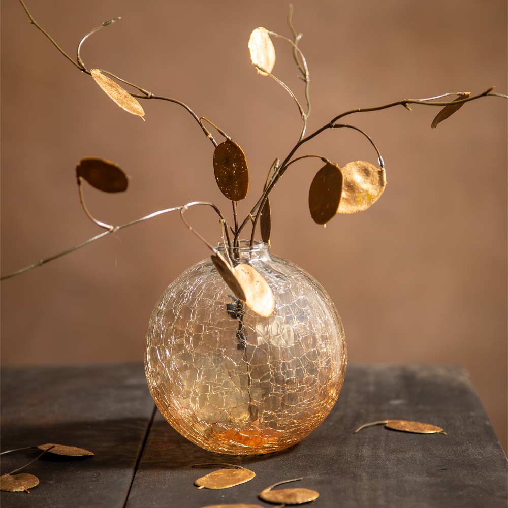A decorative metal leaf vine arrangement in a clear crackle-glass vase, set on a dark tabletop against a warm brown background.