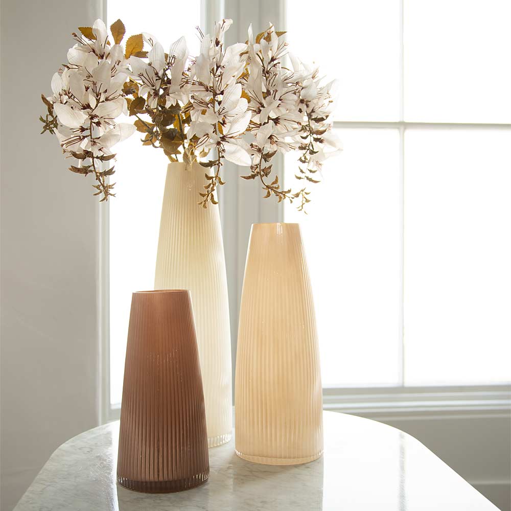 Three ribbed ceramic vases in dusty rose , cream, and light beige tones arranged on a white marble table in front of a bright window, with white faux flowers in the tallest vase.