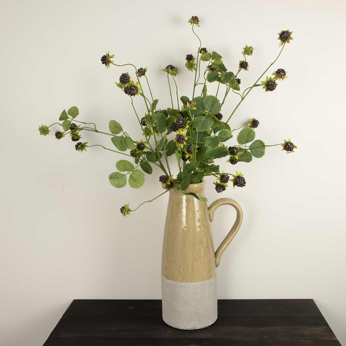 Two-Toned ceramic vase with five faux blackberry stems with green leaves on a wooden surface against a white wall