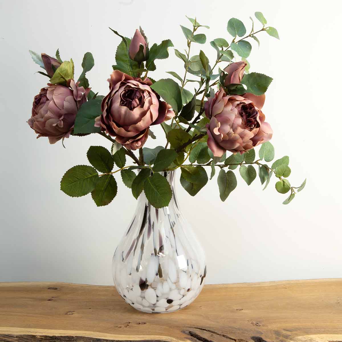 A bunch of artificial dark pink peony spray flowers arranged in a glass vase, placed on a rustic wooden surface
