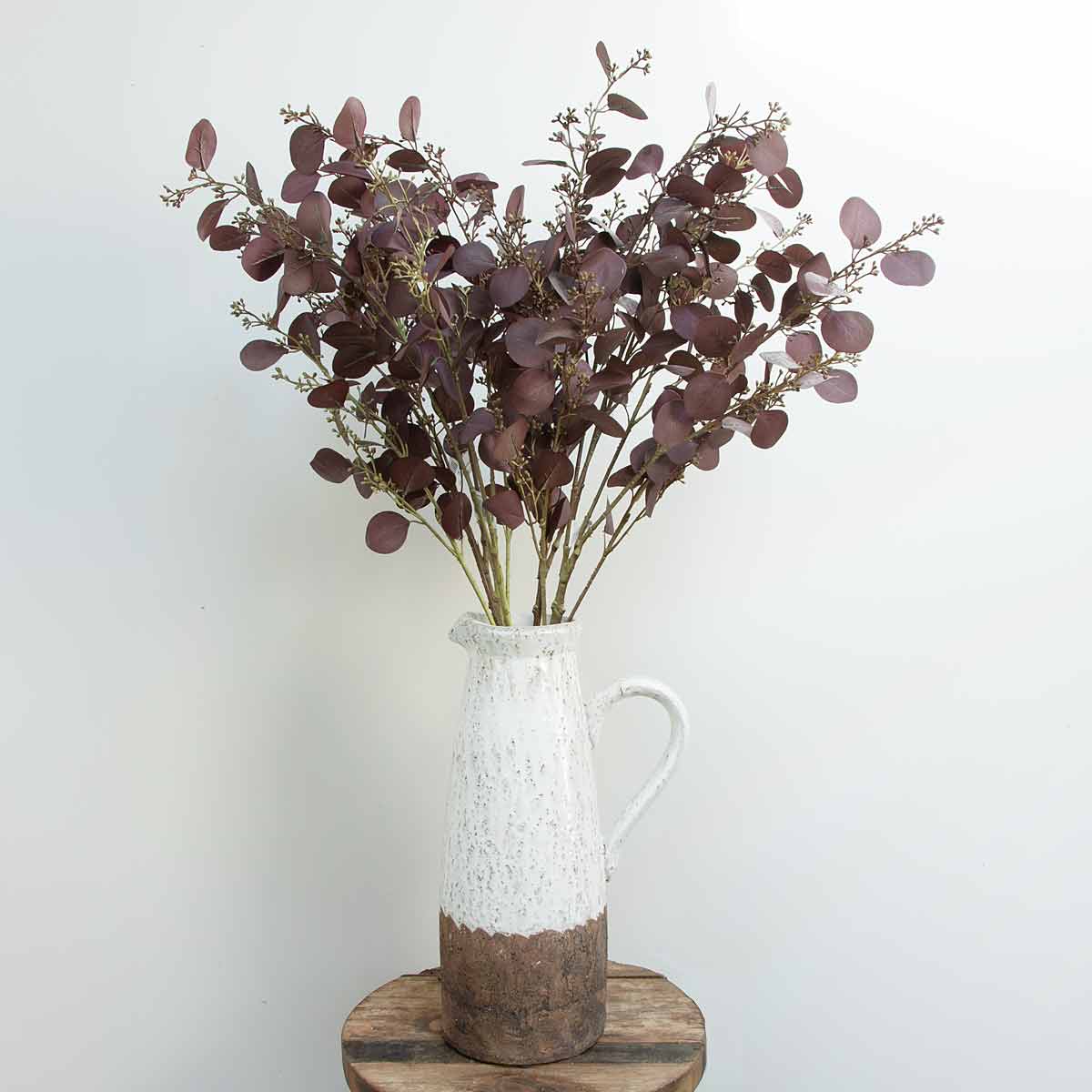 A bunch of artificial aubergine-colored eucalyptus leaves in a white vase with a brown base, placed on a wooden surface against a neutral background.