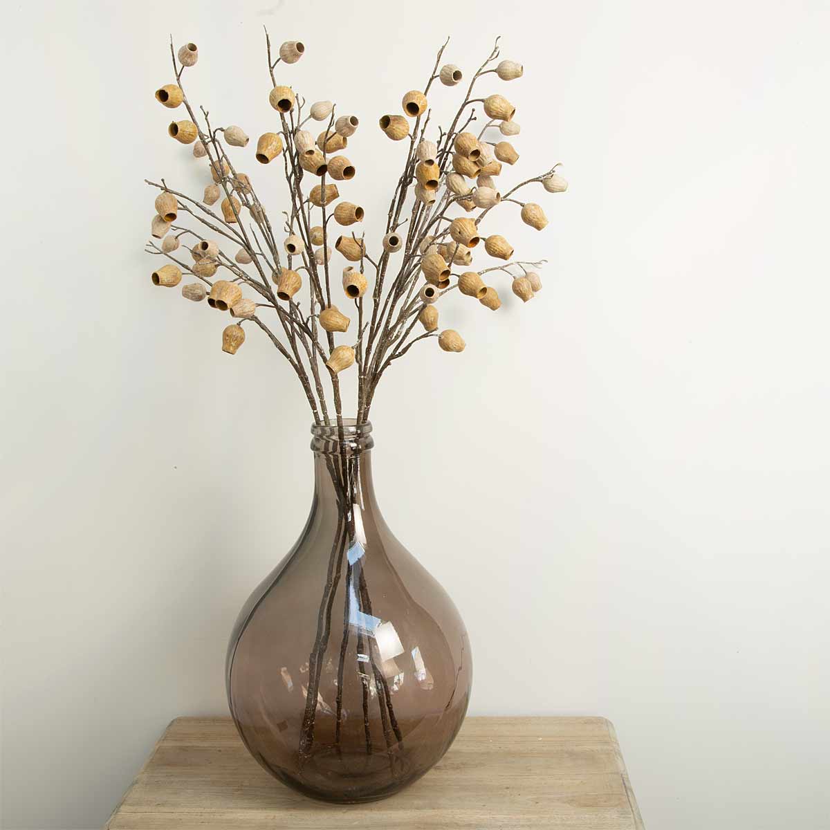 Glass vase with five faux eucalyptus pods and branches on a wooden surface against a white wall