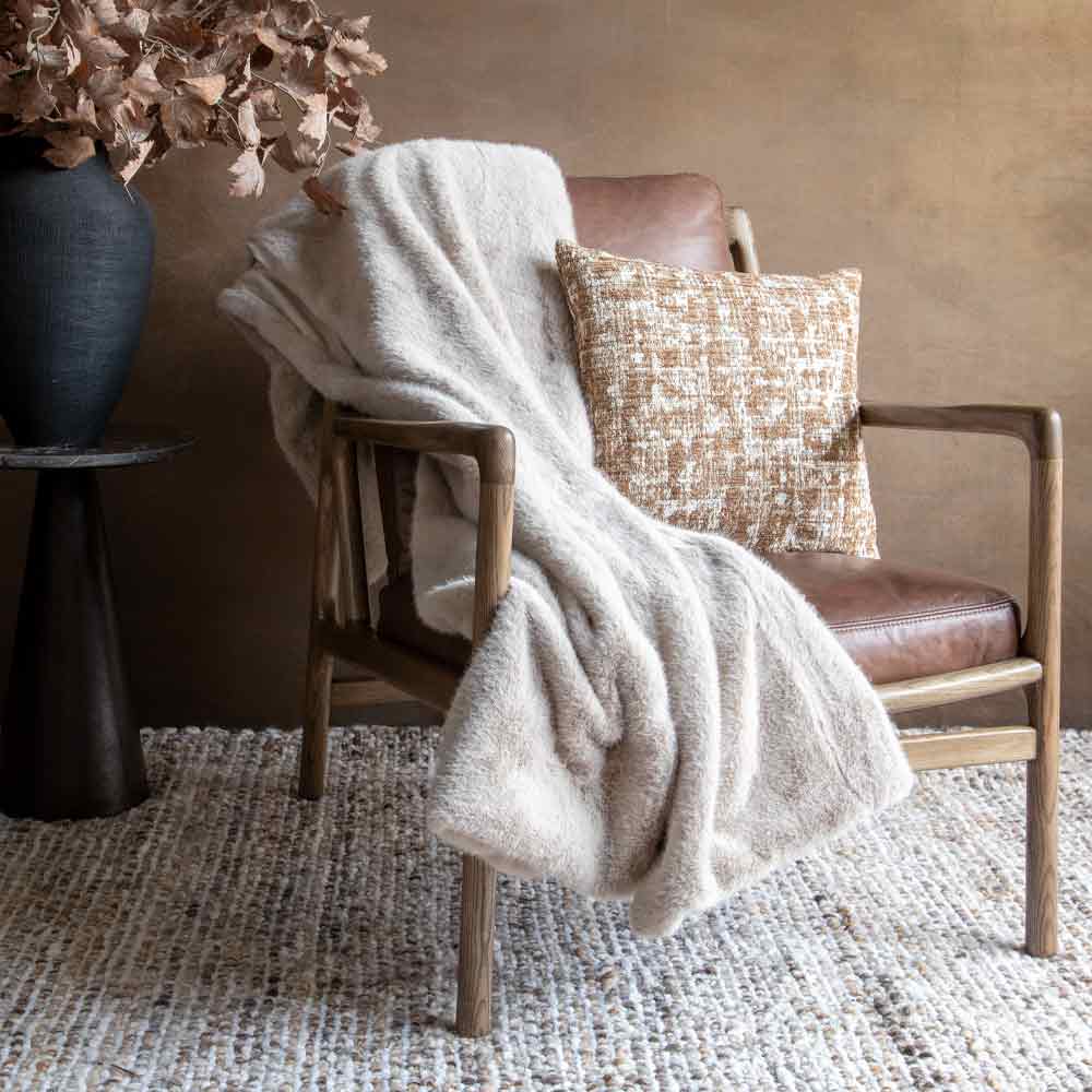 A cozy modern armchair with brown leather seating and a beige textured throw blanket draped over the arm, styled with a textured cream and brown pillowon a neutral rug against a warm brown wall.