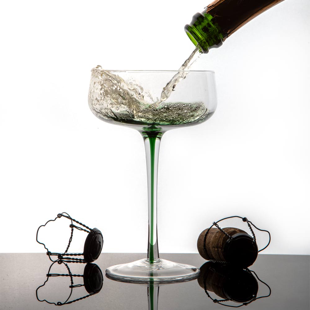 A stemmed coupe-style glass with a green-tinted stem into the bowl, shown on a reflective black surface. A green bottle is pouring a fizz iquid into the glass, creating a splash.