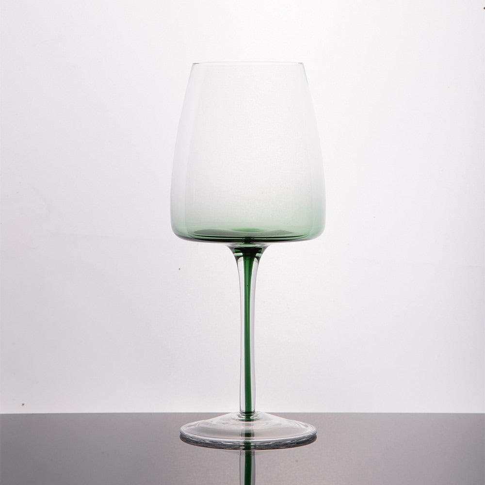 A single stemmed wine glass with a clear bowl that fades into a light green tint at the base and along the stem, standing on a dark reflective surface against a white background.