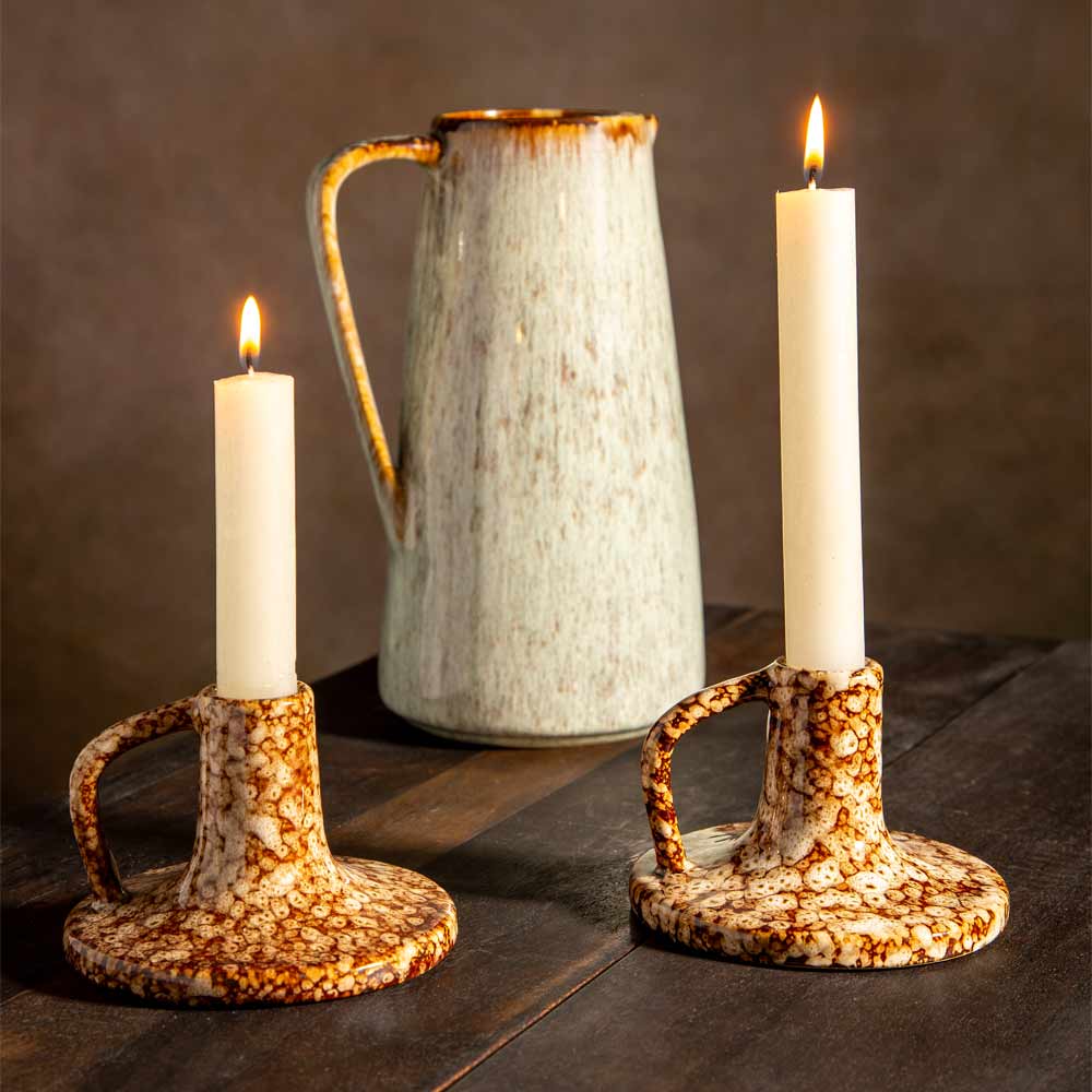 Two ceramic candlestick holders with cream dinner candles on a dark wooden table, with a speckled ceramic pitcher in the background.