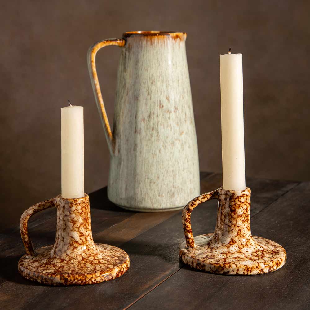 Two ceramic candlestick holders with cream and brown speckled glaze, each holding a cream dinner candle, displayed on a dark wooden table with a speckled ceramic pitcher in the background.