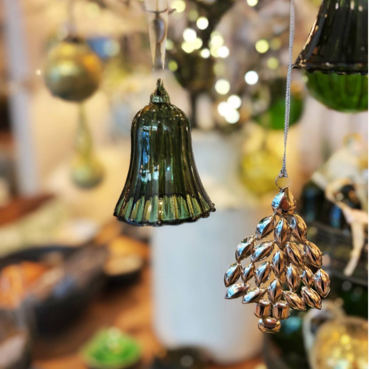 Decorative Glosille Green Glass Ornament Bell and silver tree ornament hanging against a blurred festive background.