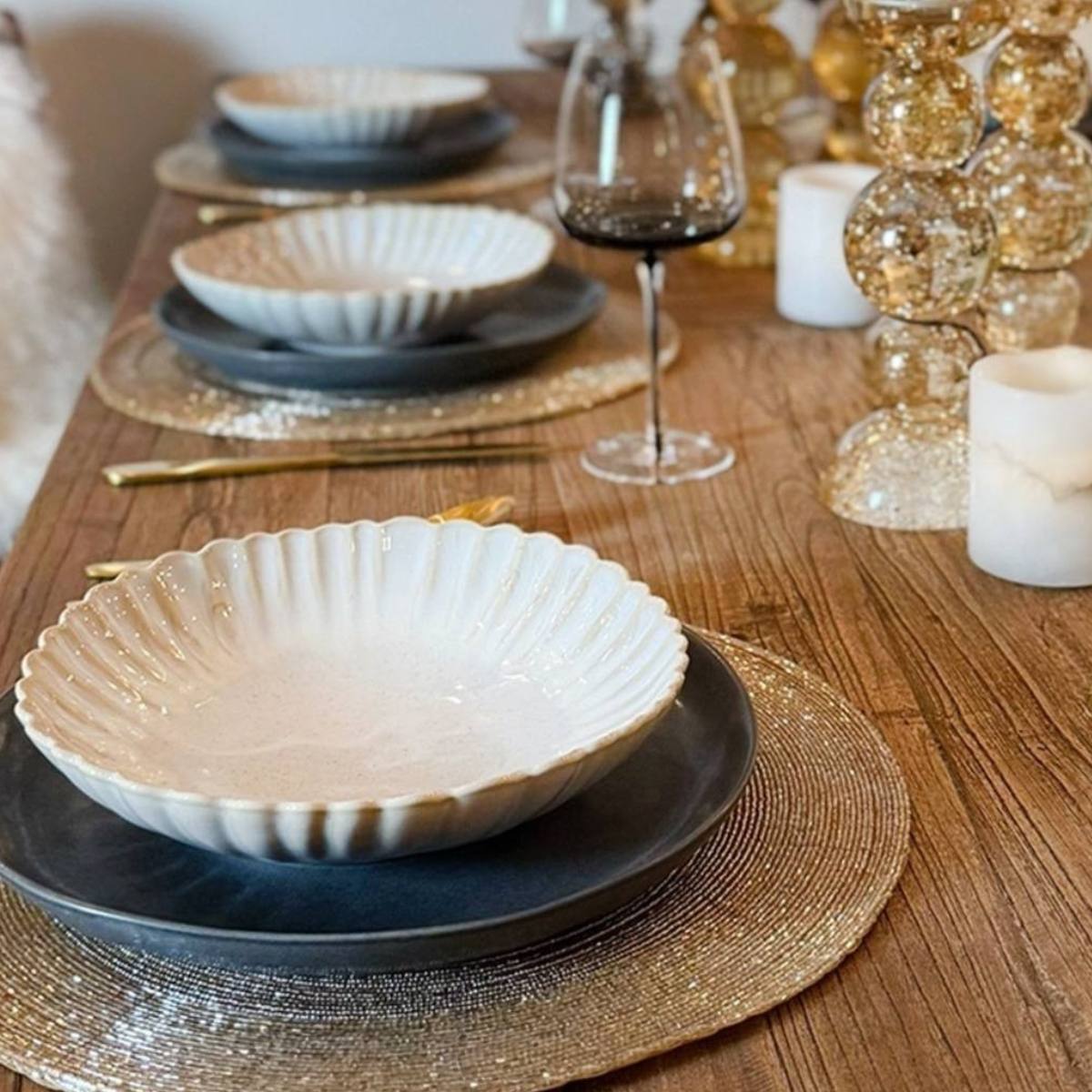 Dining table setting with ceramic bowls on Gold Woven Beaded Placemats with wine glasses and decorative items on a wooden surface.