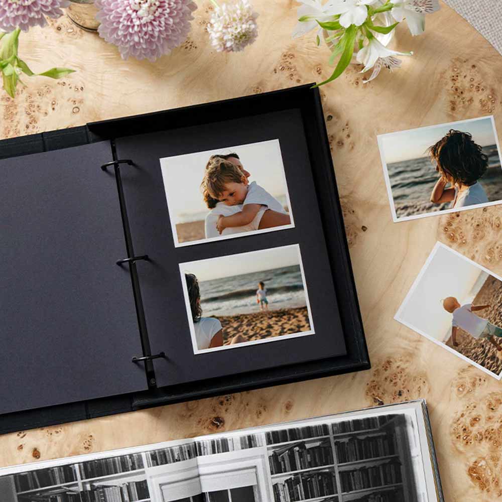 Black photo album with printed photos on a wooden surface with flowers.