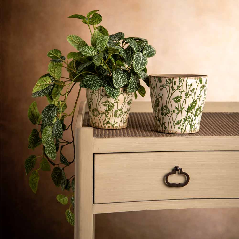 A faux potted green trailing houseplant in a cream ceramic pot with a botanical leaf pattern, displayed on a light wood side table next to another matching patterned pot against a warm brown background.