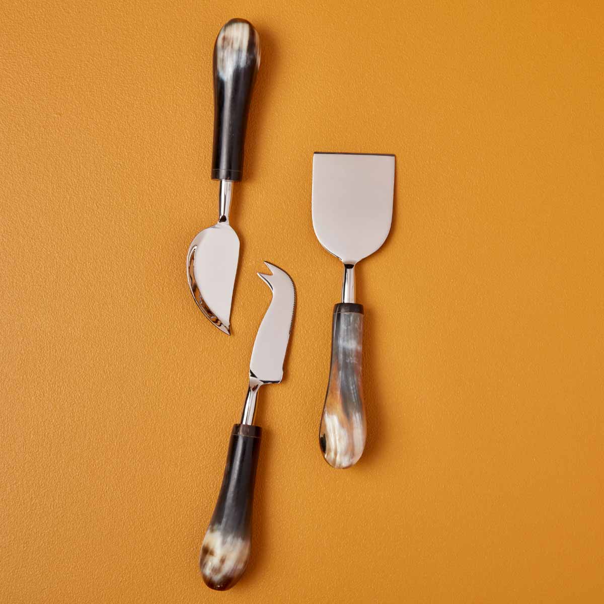 A handcrafted horn and stainless steel cheese knife set, featuring three unique utensils with ethically salvaged water buffalo horn handles, displayed on a warm ochre background.