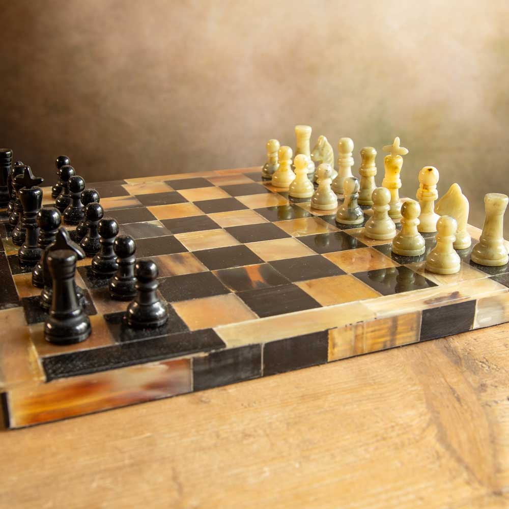 A polished chessboard made of horn with black and cream-coloured stone-like pawns and chess pieces, set on a wooden table against a warm brown background.