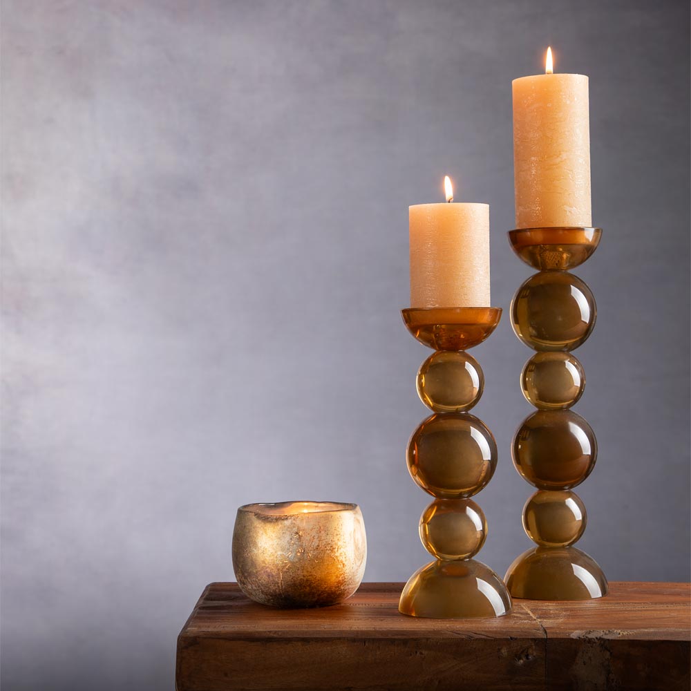 Two amber glass bubble candlestick holders with lit Honey pillar candles and a small rustic amber Votive, arranged on a rustic wooden surface against a soft gray wall background.