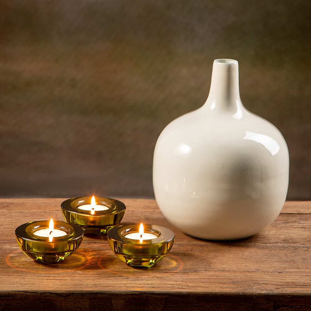 A glossy cream metal vase with a narrow neck and rounded body stands on a wooden surface next to three small lit tealight candle holders in green  glass.