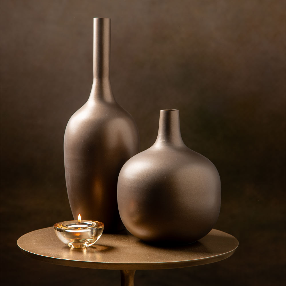 Two matte dark brown metal vases of different shapes displayed on a round wooden table, with a small lit glass candle holder in front, against a dark brown background.