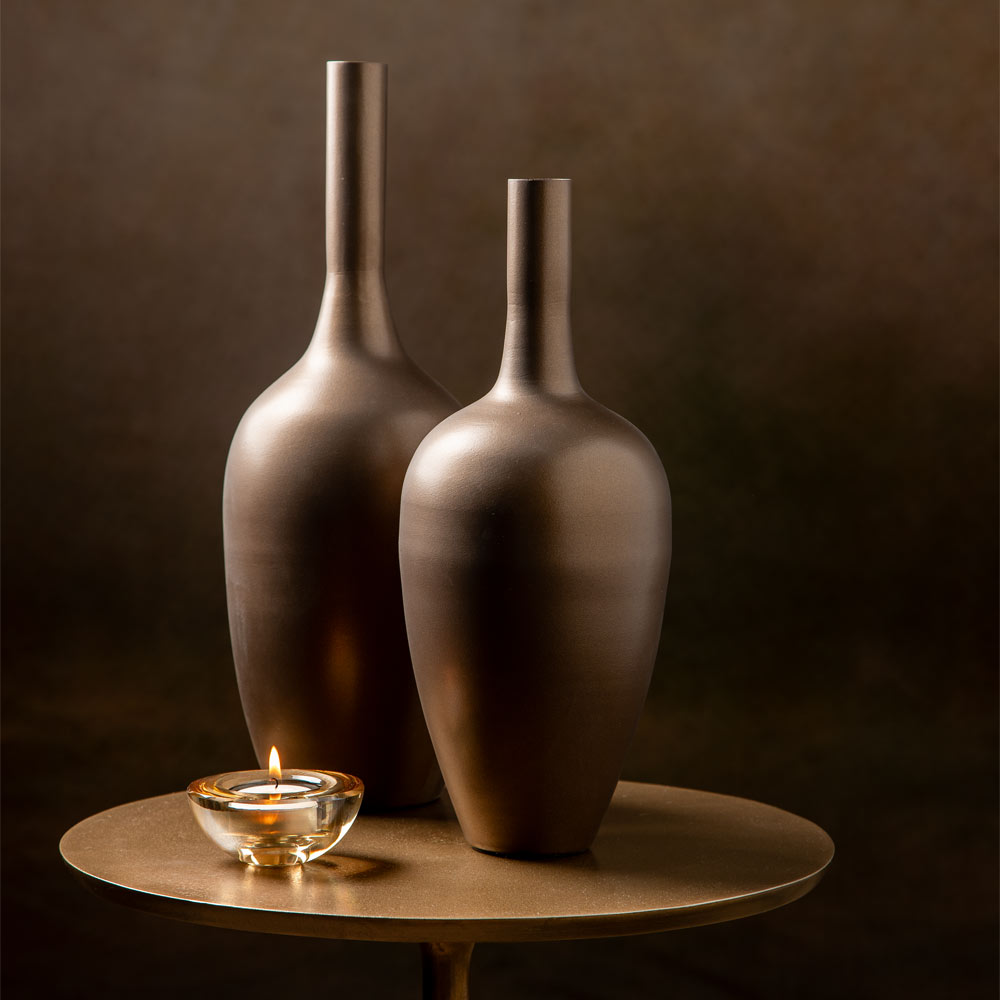 Two matte dark brown metal  vases with a minimalist bulb shape and long necks, displayed on a round bronze side table next to a lit glass tealight candle against a dark brown background.